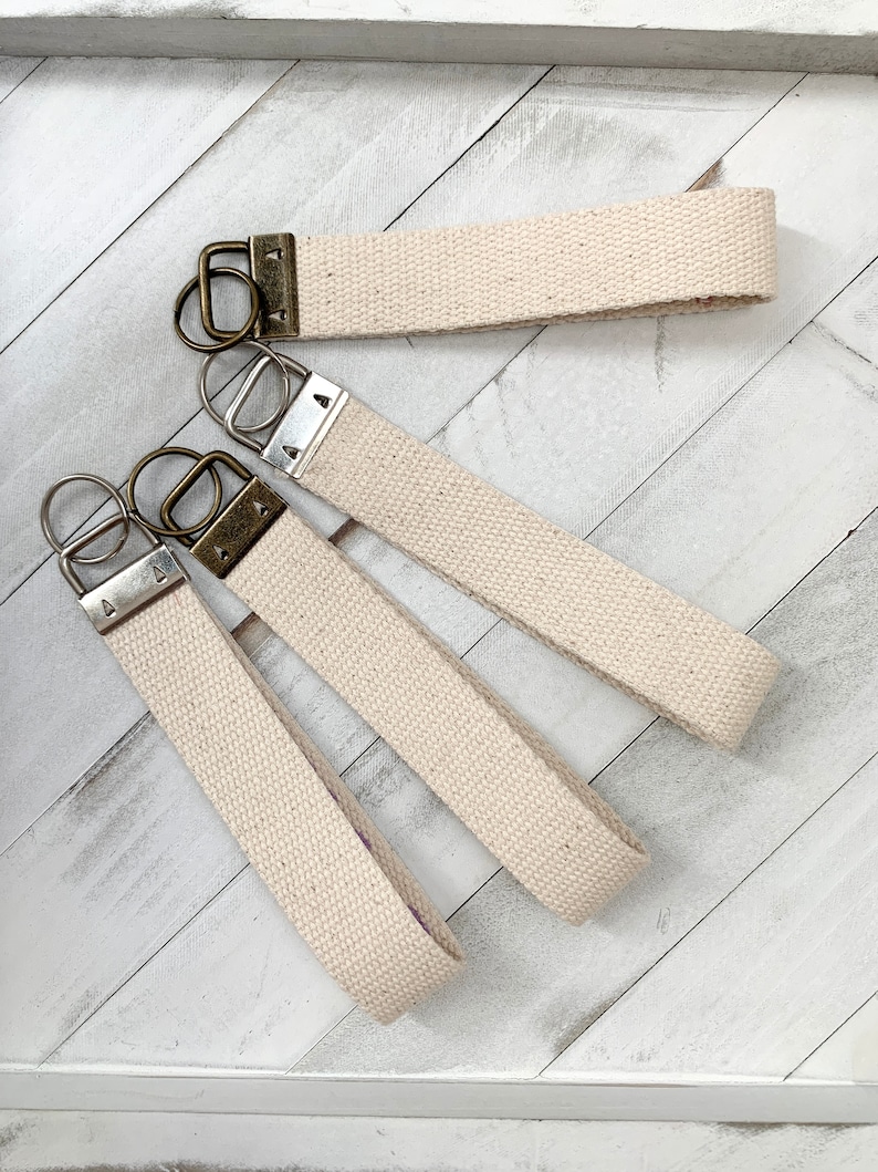 May include: Several beige canvas wrist keychains with metal hardware. The keychains have a loop design and are made of a sturdy, woven material. The metal hardware includes a key ring and a clasp.