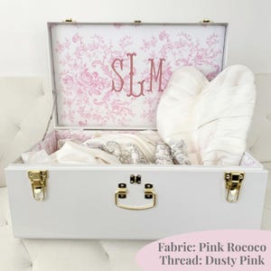 May include: A white storage trunk with gold-toned metal latches and a handle. The interior lid features a pink floral toile pattern with the embroidered monogram SLM in dusty pink thread. Inside, a white garment with silver embellishments is stored.
