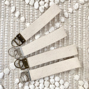 May include: Four off-white canvas wrist keychains with antique brass hardware and silver key rings. The keychains are arranged on a textured, neutral-toned surface with small, white, fabric-like accents.