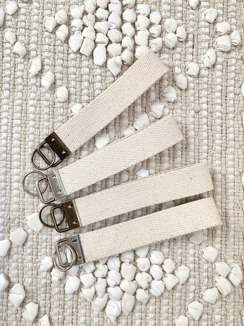 May include: Four off-white canvas wrist keychains with antique brass hardware and silver key rings. The keychains are arranged on a textured, neutral-toned surface with small, white, fabric-like accents.