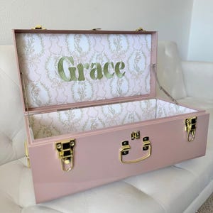 Könnte beinhalten: Eine staubrosa Metall-Aufbewahrungstruhe mit goldfarbenen Verschlüssen und einem Griff. Das Innere ist mit weißem Papier mit hellrosa Blumen- und Schleifenmuster ausgekleidet, wobei der Name Grace in grünem Garn auf dem Deckel gestickt ist.