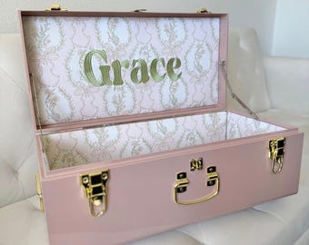 May include: A dusty pink metal storage trunk with gold-toned latches and a handle. The interior is lined with white paper featuring a light pink floral and ribbon pattern, with the name Grace embroidered in green thread on the lid.