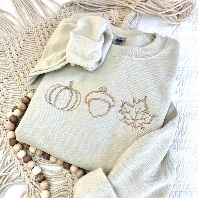 May include: A light beige sweatshirt with an embroidered design of a pumpkin, acorn, and maple leaf. The design is in a brown color.