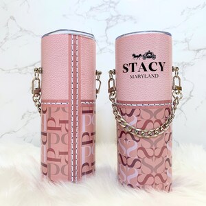 May include: Two pink stainless steel tumblers with a chain handle. One tumbler has a pink and white monogram pattern and the other has the name "Stacy Maryland" printed on it.