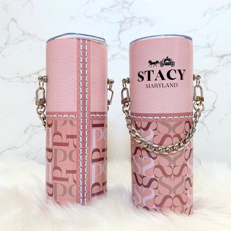 May include: Two pink stainless steel tumblers with a chain handle. One tumbler has a pink and white monogram pattern and the other has the name "Stacy Maryland" printed on it.