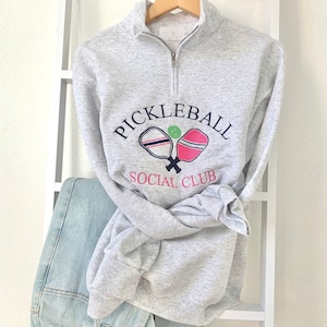 May include: A gray sweatshirt with a white and navy blue graphic of two pickleball paddles and a green ball. The text "Pickleball Social Club" is printed in navy blue.