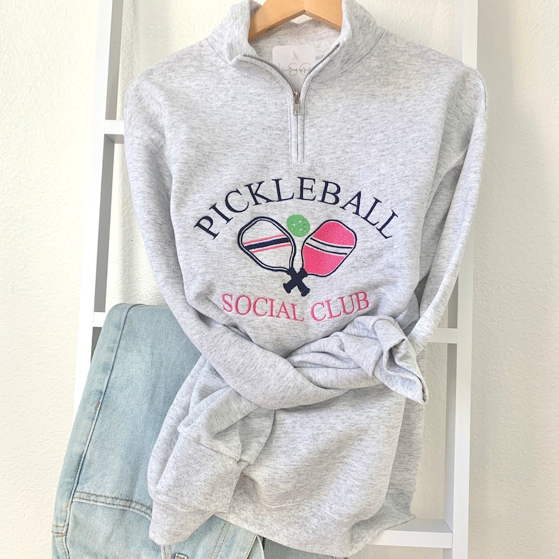 May include: A gray sweatshirt with a white and navy blue graphic of two pickleball paddles and a green ball. The text "Pickleball Social Club" is printed in navy blue.