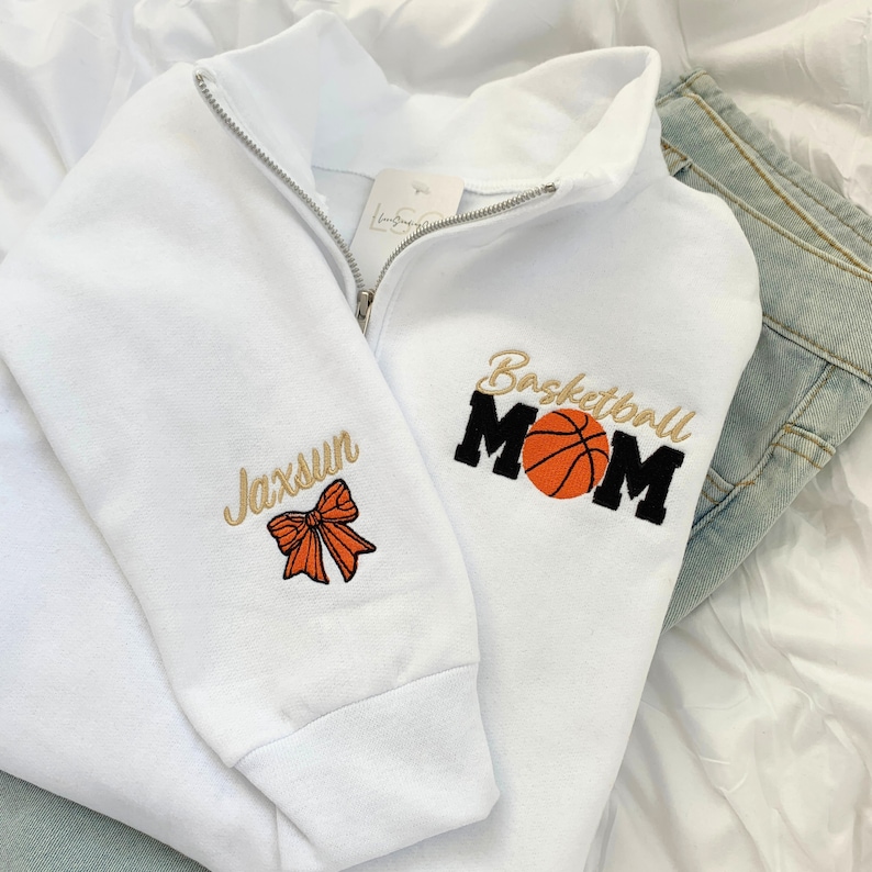 May include: White half-zip sweatshirt with embroidered designs.  One sleeve shows 'Jaxsun' in script with a bow. The other side displays 'Basketball MOM' in bold text with an orange basketball graphic. The sweatshirt is shown folded on a light surface with a pair of light blue jeans.