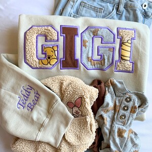 May include: A beige sweatshirt with the word "GIGI" embroidered in purple and brown letters. The sweatshirt has a fuzzy texture and the letters are decorated with Winnie the Pooh characters. The sweatshirt also has the words "Teddy Bear" embroidered on the sleeve.
