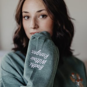 May include: A heathered forest green long-sleeve sweatshirt featuring white embroidered text on the sleeve that reads Bodhi Willow Ranger. The garment includes a small, colorful embroidered graphic on the chest area, showcasing a detailed floral design.