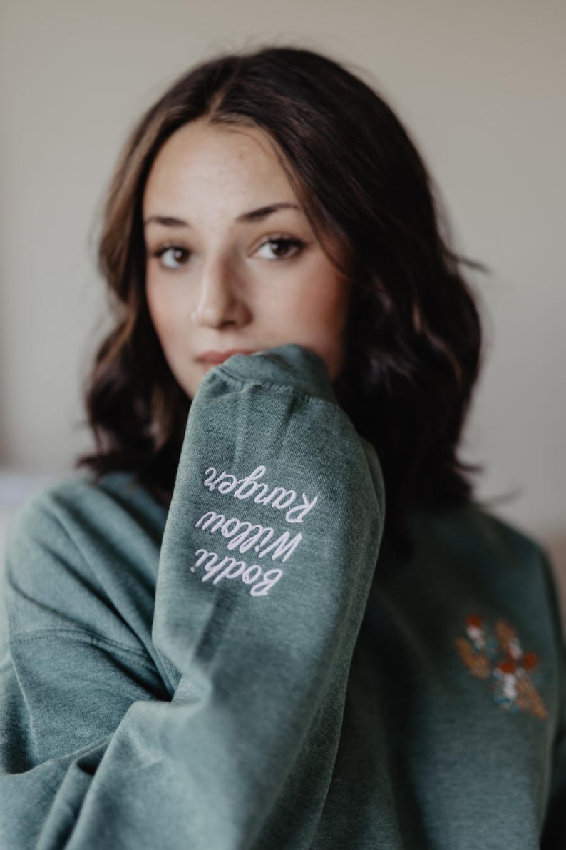 May include: A heathered forest green long-sleeve sweatshirt featuring white embroidered text on the sleeve that reads Bodhi Willow Ranger. The garment includes a small, colorful embroidered graphic on the chest area, showcasing a detailed floral design.