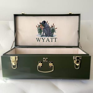 May include: An olive green metal storage box with gold-toned latches and a central handle. The interior lid features an embroidered graphic of a dog sitting among cattails with the name WYATT stitched in black lettering below the image.