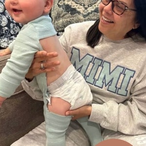 May include: A woman wearing a gray sweatshirt with the word "MIMI" printed on it is holding a baby wearing a light blue onesie and a diaper. The baby is standing on the woman's lap. The image is captioned "KEEPSAKE GIFT".