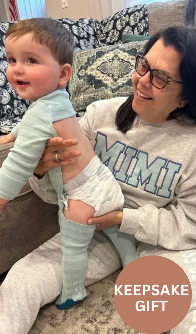May include: A woman wearing a gray sweatshirt with the word "MIMI" printed on it is holding a baby wearing a light blue onesie and a diaper. The baby is standing on the woman's lap. The image is captioned "KEEPSAKE GIFT".