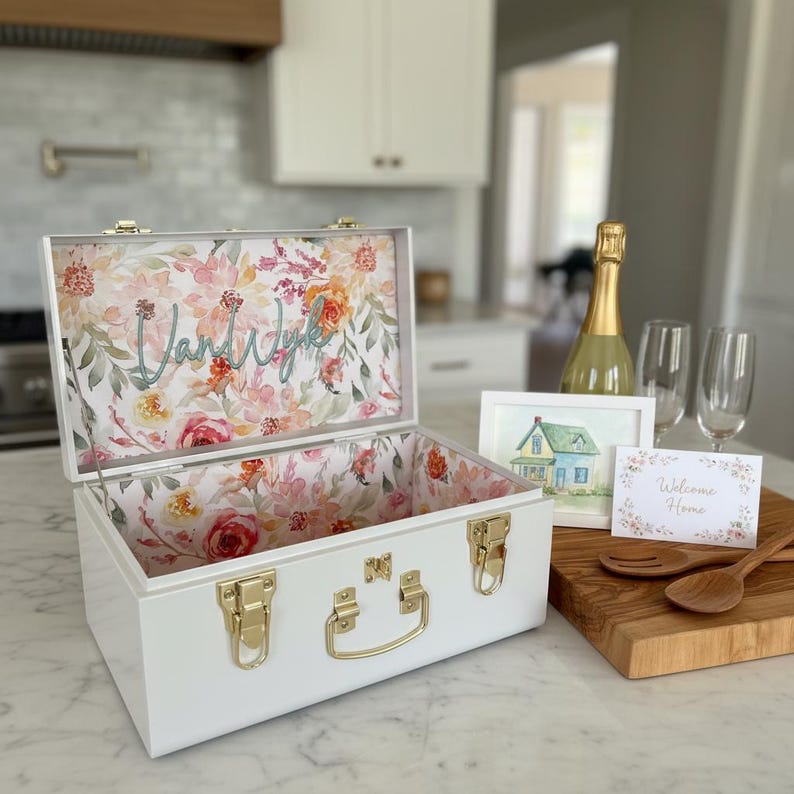 May include: White decorative box with gold hardware and a floral interior. The inside lid features the name "Vand Wyk" in cursive. A watercolor painting of a house and a "Welcome Home" card are on a wooden cutting board.