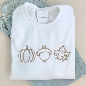 May include: White sweatshirt with a brown embroidered pumpkin, acorn, and maple leaf design. The text "Cozy Fall Styles" is printed below the sweatshirt.