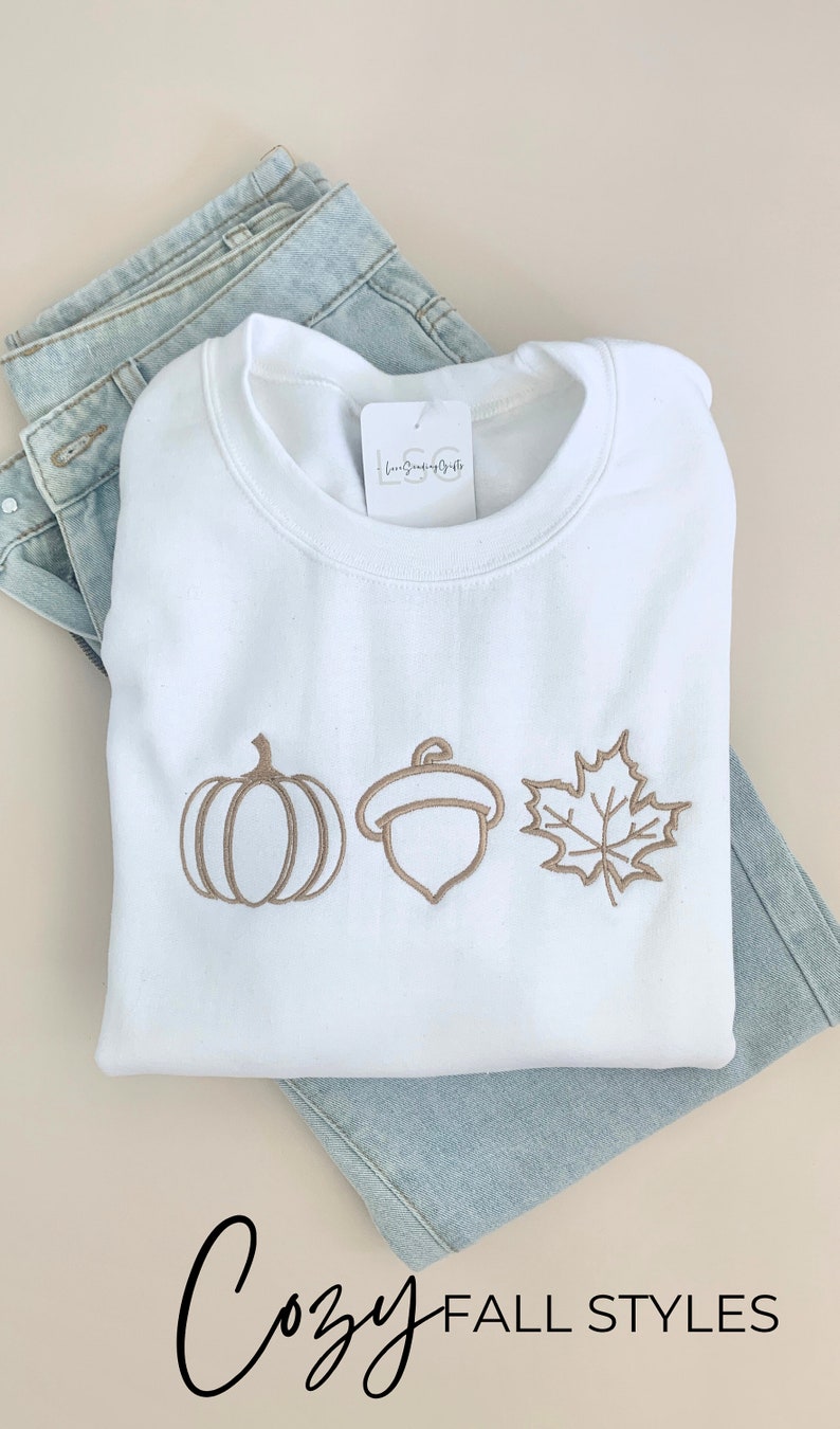 May include: White sweatshirt with a brown embroidered pumpkin, acorn, and maple leaf design. The text "Cozy Fall Styles" is printed below the sweatshirt.