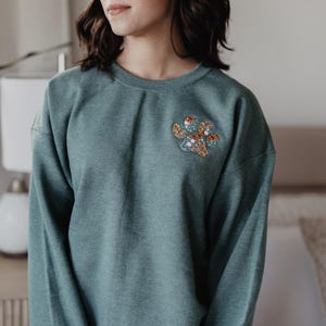 May include: A teal crewneck sweatshirt featuring an embroidered floral paw print design on the left chest. The right sleeve cuff displays the white embroidered text Bodhi Willow Ranger in a script font. The garment has long sleeves.