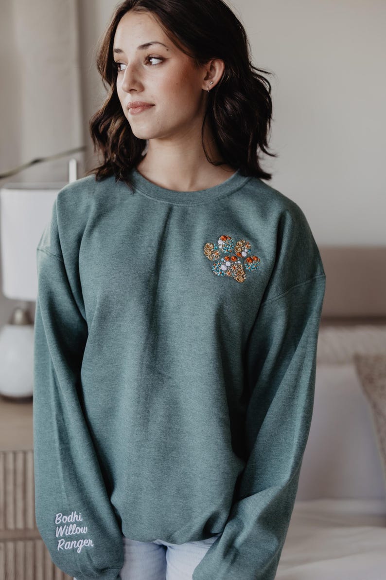 May include: A teal crewneck sweatshirt featuring an embroidered floral paw print design on the left chest. The right sleeve cuff displays the white embroidered text Bodhi Willow Ranger in a script font. The garment has long sleeves.