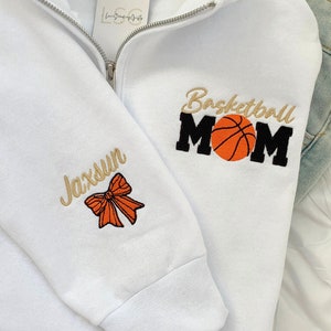 May include: White sweatshirt with a zipper and embroidered text that says "Basketball Mom" with a basketball graphic. The left sleeve has a gold embroidered bow and the text "Jaxsun".