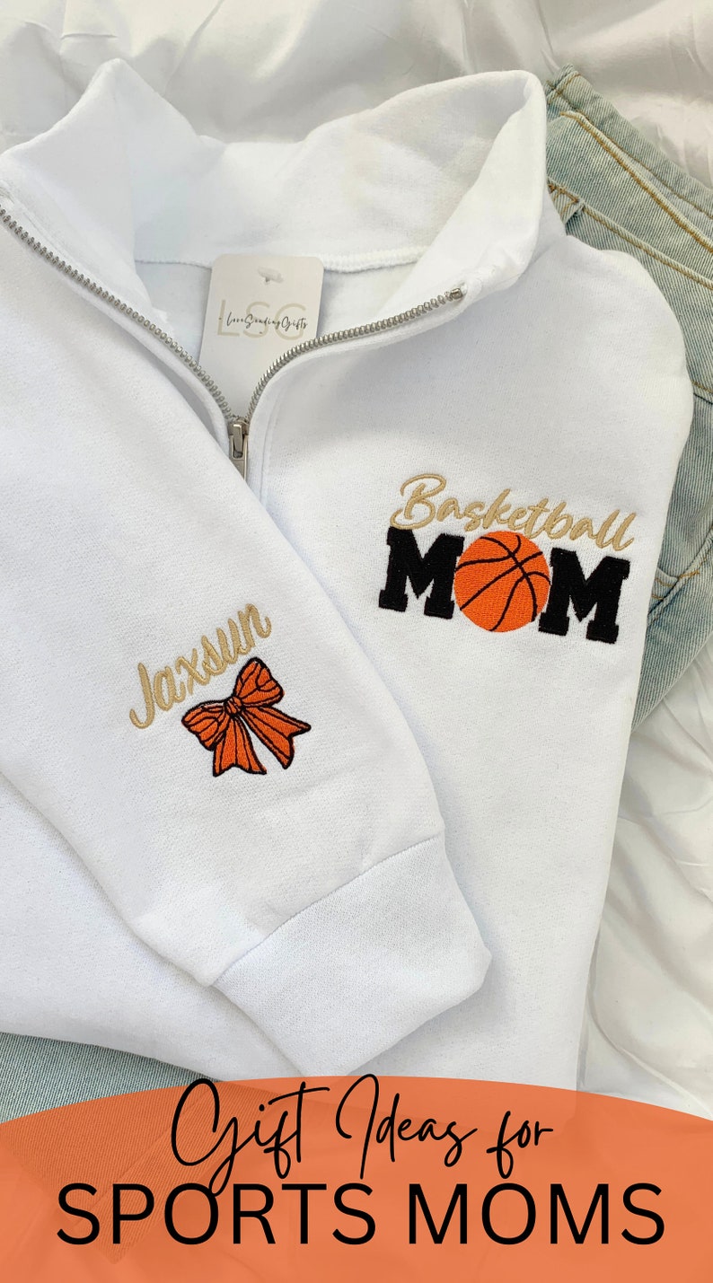 May include: White sweatshirt with a zipper and embroidered text that says "Basketball Mom" with a basketball graphic. The left sleeve has a gold embroidered bow and the text "Jaxsun".