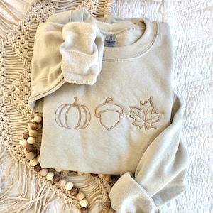 May include: A light beige sweatshirt with an embroidered design of a pumpkin, acorn, and maple leaf. The sweatshirt is folded and lying on a white surface.