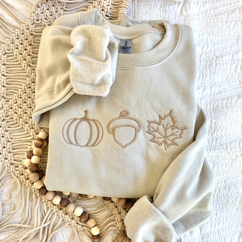 May include: A light beige sweatshirt with an embroidered design of a pumpkin, acorn, and maple leaf. The sweatshirt is folded and lying on a white surface.