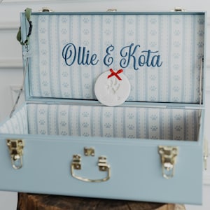 May include: A light blue keepsake chest with gold-tone metal latches and a handle. The interior is lined with light blue and white striped fabric featuring paw prints. The text Ollie & Kota is embroidered in blue, with a white clay paw print ornament.
