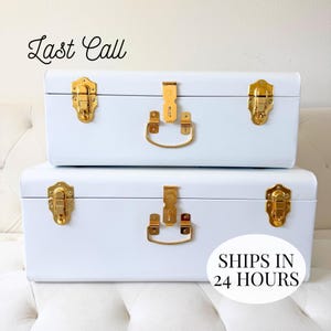 Puede incluir: Dos baúles de almacenamiento rectangulares de metal blanco apilados con cierres y asas dorados. El baúl superior presenta el texto Last Call en letra cursiva negra, y el inferior incluye un gráfico circular blanco con el texto SHIPS IN 24 HOURS.
