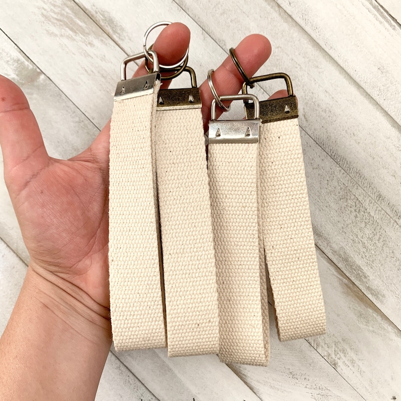 May include: Four cream-colored canvas keychains with metal hardware. The keychains have a rectangular shape and are held together by metal rings. The hardware has a silver and bronze finish. The keychains are displayed against a white wooden background.