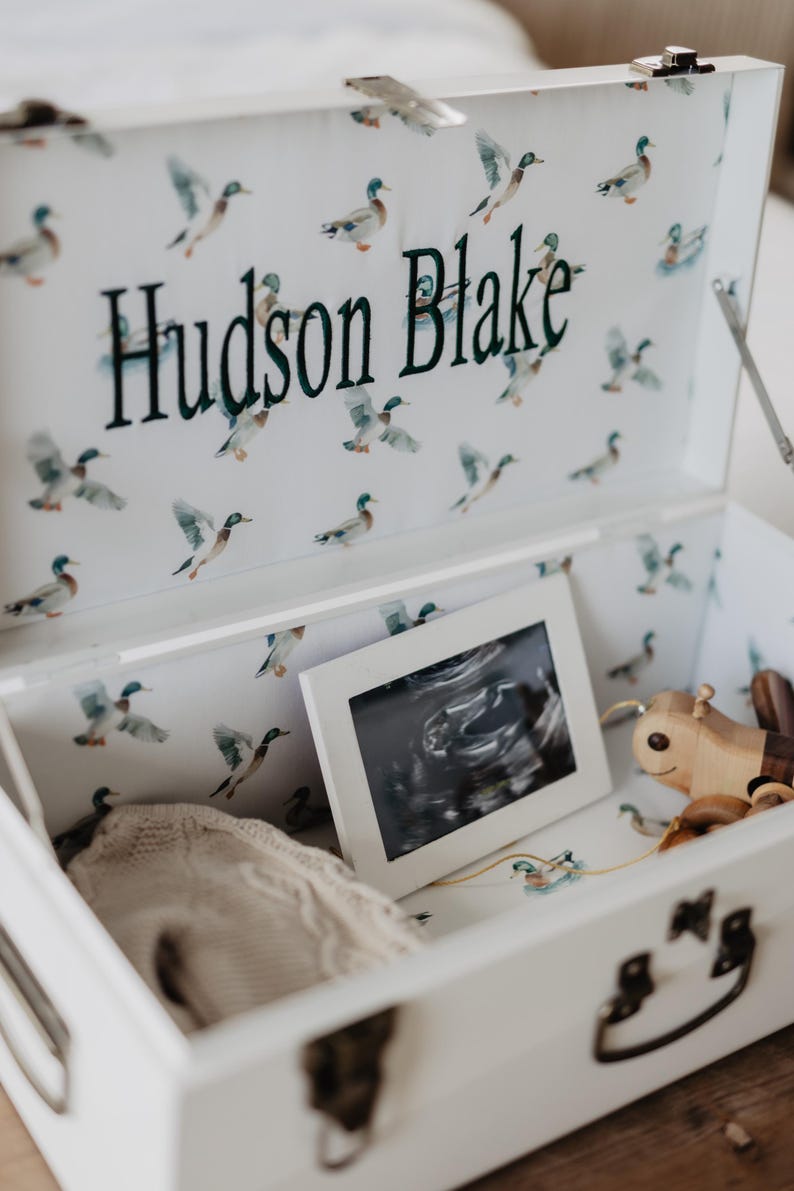 A white trunk with a pattern of ducks and the name "Hudson Blake" printed on it, containing a framed photograph and a wooden toy.