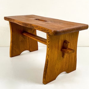 Könnte beinhalten: Ein rustikaler Holzhocker mit schlichtem Design. Der Hocker ist aus Naturholz mit einer warmen braunen Farbe gefertigt. Er hat eine rechteckige Oberseite mit einem kleinen Ausschnitt und einen einzigen horizontalen Trägerbalken. Die Beine sind leicht gebogen und haben ein einzigartiges, handgefertigtes Aussehen.