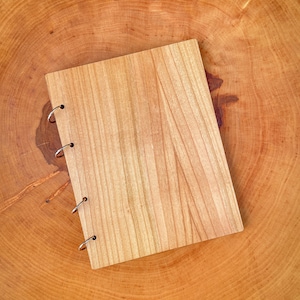 Può includere: Un taccuino di legno con una rilegatura ad anelli. Il taccuino è di colore marrone chiaro con una venatura del legno. Il taccuino è appoggiato su una superficie di legno.