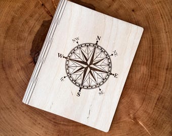 May include: A light-colored wooden notebook with a compass rose design etched on the cover. The compass rose features directional indicators such as North, South, East, and West. The notebook is resting on a wooden surface.