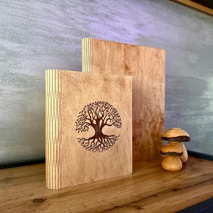 Può includere: Due fermalibri in legno e una statuina a forma di fungo. I fermalibri sono in legno chiaro, con un disegno dell'albero della vita inciso su uno. La statuina a forma di fungo è marrone chiaro con un cappello marrone più scuro.