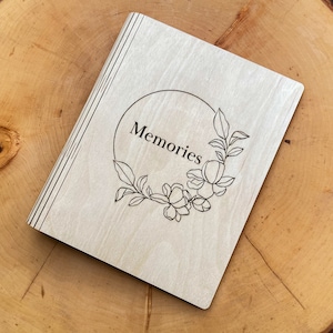Può includere: Un album fotografico in legno chiaro con la scritta "Memories" all'interno di una ghirlanda floreale. L'album è chiuso e presenta una serie di linee verticali sul dorso. L'album è su una superficie di legno.