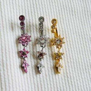 May include: Three belly button rings featuring dangling flower-shaped charms adorned with sparkling crystals. One ring is silver-toned with pink stones, one is silver-toned with clear stones, and one is gold-toned with clear stones.