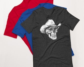 Puede incluir: Tres camisetas de manga corta en rojo, azul y gris antracita. La camiseta gris antracita presenta una ilustración detallada en blanco y negro de una calavera con un sombrero de vaquero. El sombrero tiene un diseño de calavera y huesos cruzados.