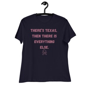 May include: Navy blue t-shirt with the text "THERE'S TEXAS, THEN THERE IS EVERYTHING ELSE." in pink lettering. A pink Texas outline with crossed bones is below the text. The t-shirt is hanging on a black hanger.