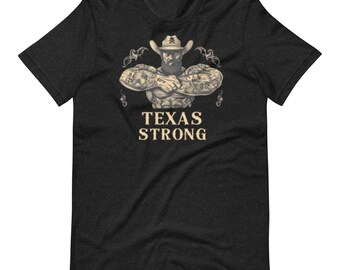 Puede incluir: Camiseta gris oscuro con una ilustración de un vaquero musculoso con tatuajes y sombrero. El texto "TEXAS STRONG" está impreso debajo en una fuente serif. La camiseta está hecha de un material suave.