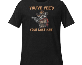 Puede incluir: Camiseta gris oscuro con un mapache de dibujos animados con sombrero y bufanda de vaquero, sosteniendo una pistola. El texto de arriba dice "YOU'VE YEE'D" y abajo "YOUR LAST HAW".