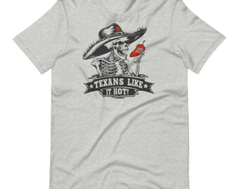 Puede incluir: Camiseta gris claro con un esqueleto con sombrero y un chile rojo. El diseño incluye el texto "TEXANS LIKE IT HOT!" en una pancarta. La camiseta es de cuello redondo y manga corta.