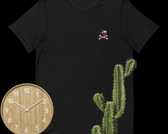 Op de afbeelding: Zwart t-shirt met een geborduurde Texas-vlag en gekruiste botten. Een houten klok en een cactus staan op de achtergrond.