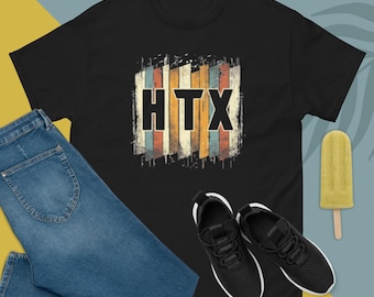 Puede incluir: Camiseta negra con la palabra "HTX" en un diseño retro. El gráfico presenta rayas verticales en varios colores. La camiseta se combina con vaqueros azules y zapatillas negras. Un helado amarillo también está en la imagen.