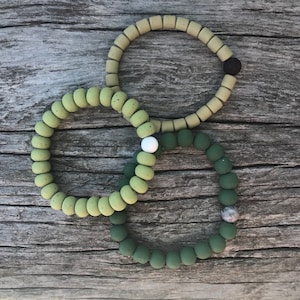 May include: Three silicone bracelets in shades of green. The top bracelet is light green with a black bead. The middle bracelet is a darker shade of green with a white bead. The bottom bracelet is a deep green with a gray bead.