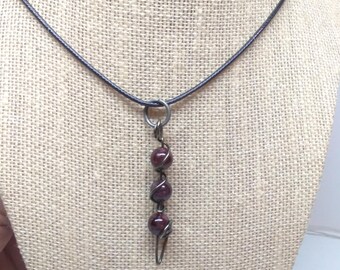 May include: A necklace featuring a dark metal wire-wrapped pendant with three round, deep red garnet-colored beads suspended vertically from a thin black cord. The pendant is attached to the cord by a simple dark metal jump ring.