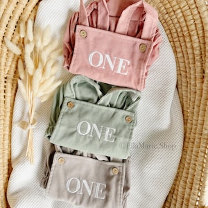May include: Three corduroy baby rompers in pink, sage green, and taupe, each embroidered with the word "ONE" in white. The rompers have wooden buttons and adjustable straps, displayed in a woven basket.