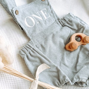 May include: Pale green corduroy romper with the word "ONE" in white. The romper features wooden button fastenings and suspender straps. A small wooden car toy is placed on the romper. The romper is on a white textured surface.