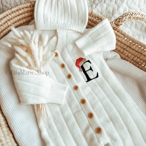 Könnte beinhalten: Weißes gestricktes Baby-Outfit mit passender Mütze. Das Outfit hat einen Knopfverschluss und einen festlichen gestickten Buchstaben "E" mit einer Weihnachtsmütze oben drauf.