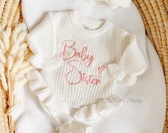 May include: A white knit baby outfit set, including a headband with a bow, a long-sleeved onesie with the words "Baby Sister" embroidered in pink, and matching booties. The set is displayed on a white blanket.